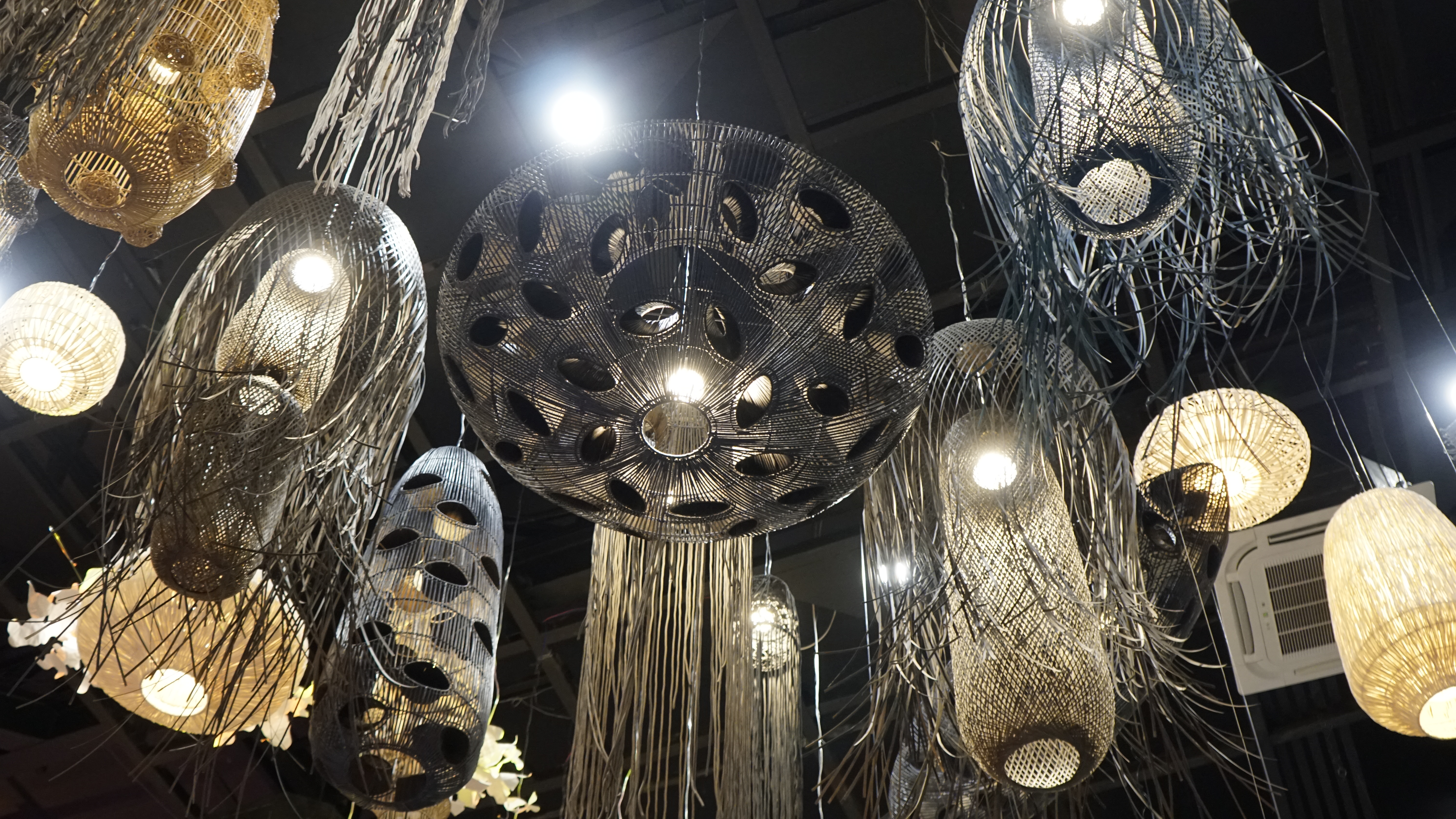 A low-angle view looking up at a cluster of intricate hanging pendant lights made from dark and light natural fibers with open-weave patterns.