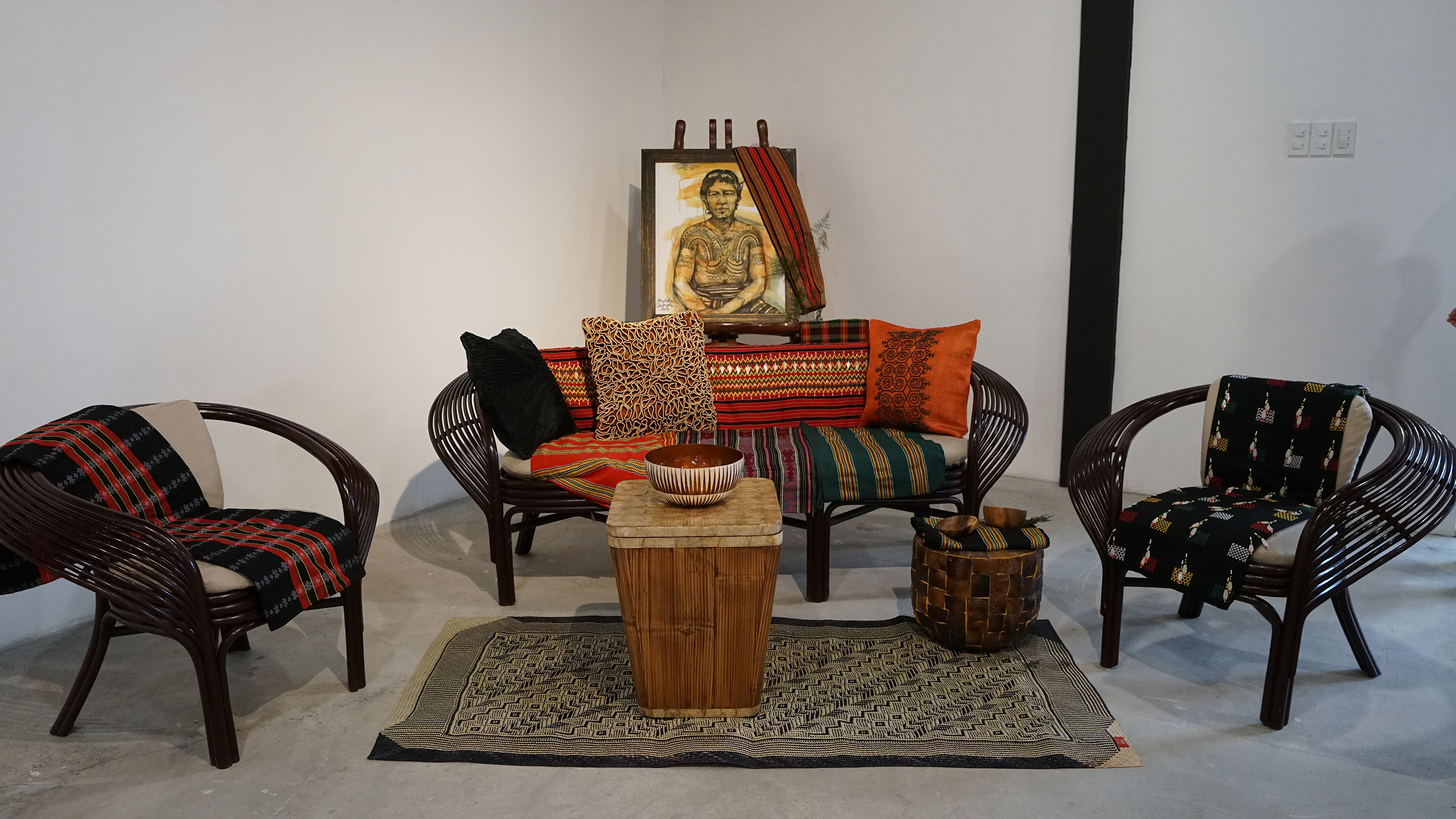 A cozy sitting area with dark rattan chairs, colorful woven throw pillows, and a painting on an easel.