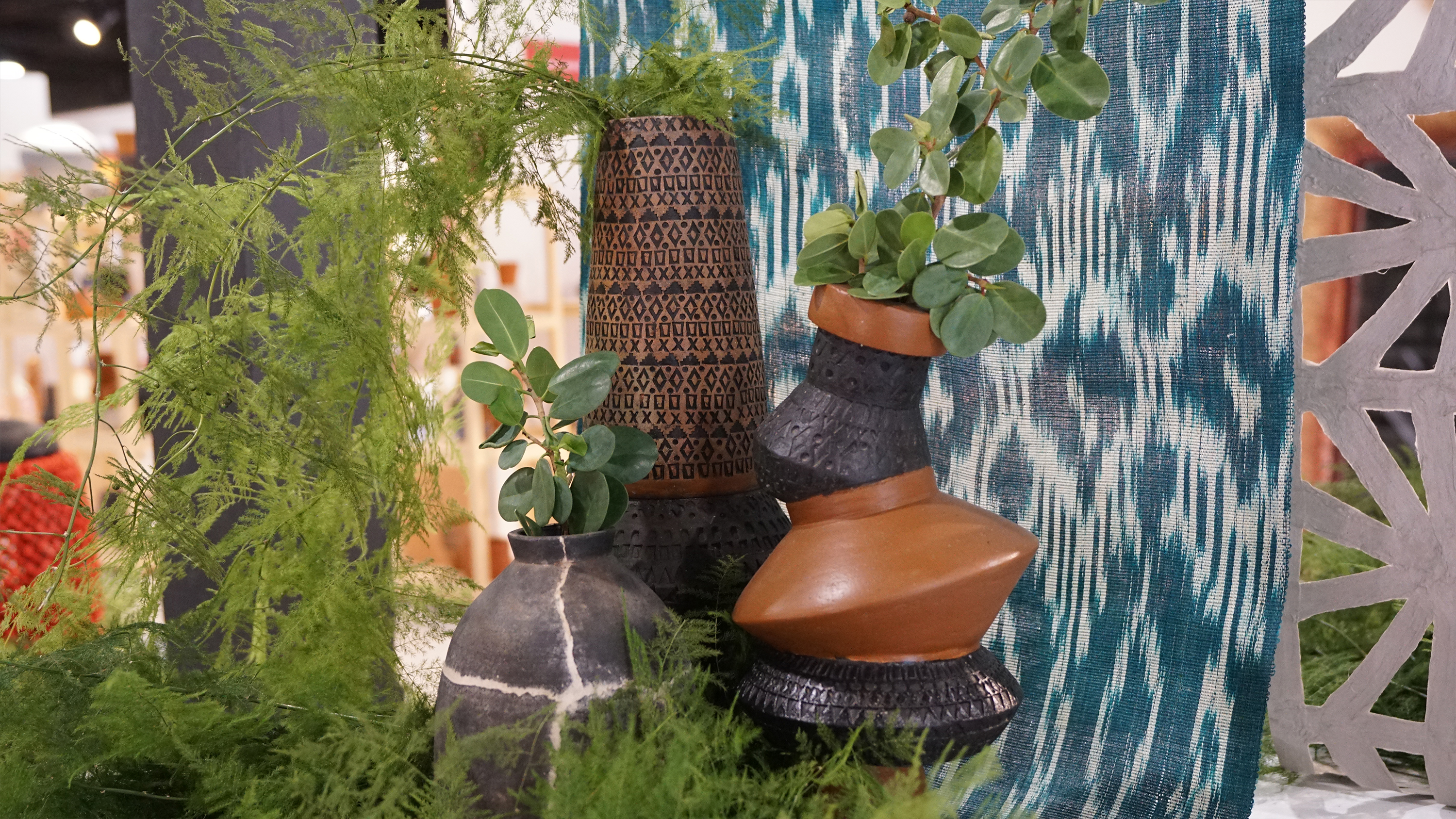 Tall brown and black patterned vases holding green plants against a blue woven fabric background.